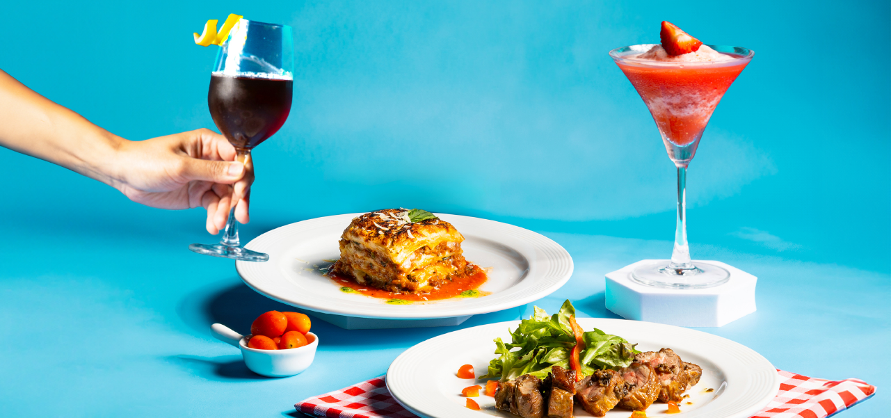 A table setting with a lasagna dish, a salad with meat skewers, a hand holding a glass with a drink, and a strawberry-topped frozen drink. A small bowl of cherry tomatoes is also present.